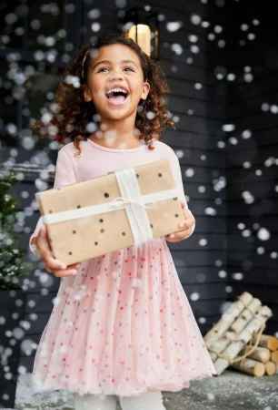 A cheerful young girl with a present stands outside in a pink dress as snow falls, capturing a happy holiday moment with festive decorations.