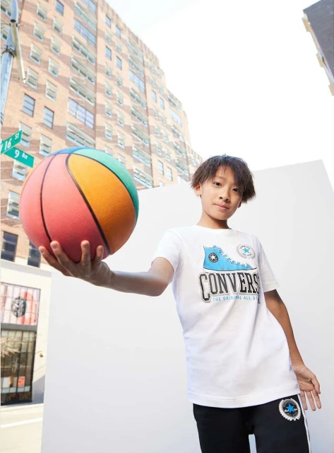 Young boy in Converse tee holds a colorful basketball in an urban setting with brick buildings and white walls creating a bright, sporty look.