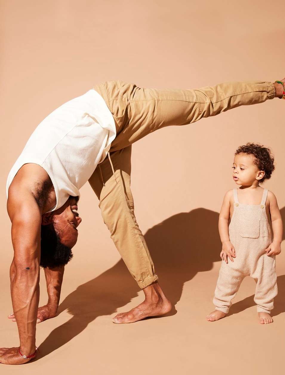 Man performing a yoga stretch with a toddler standing nearby against a warm neutral backdrop