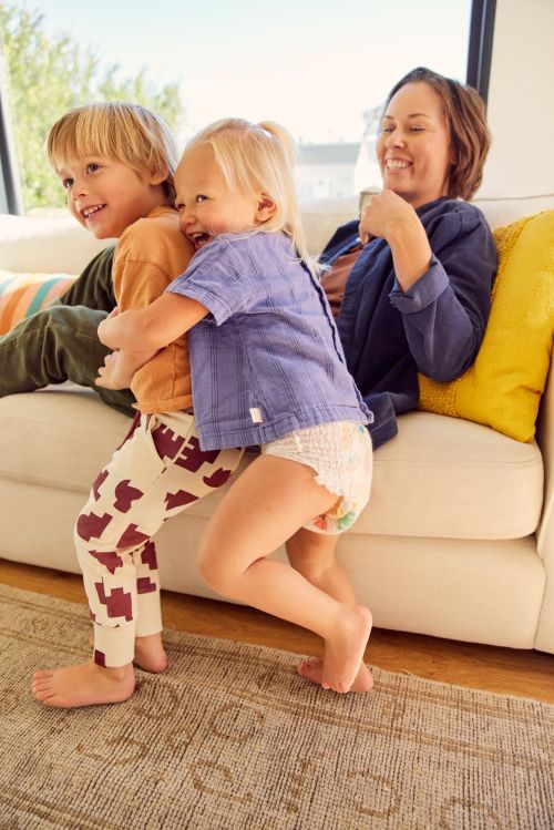 Two young children hugging while sitting on a white couch next to an adult woman in a casual blue top smiling indoors.