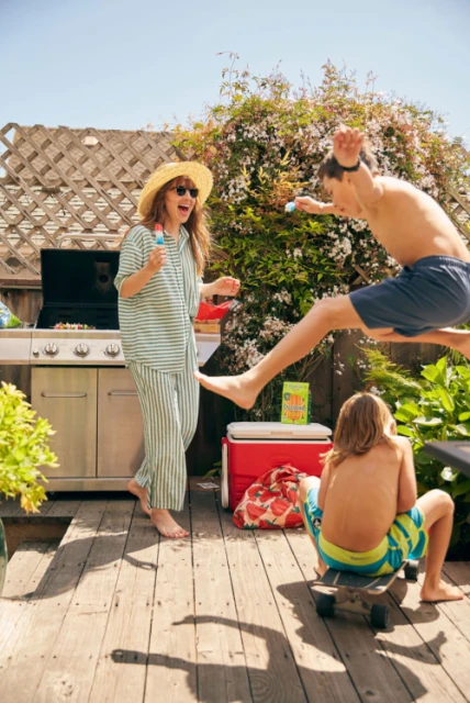 Family fun on a sunny deck with a grill, cooler, and foliage: a woman in pajamas, a jumping boy, and a child on a skateboard.