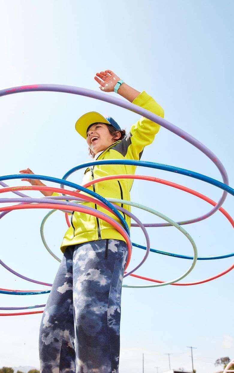 A young child in a yellow jacket smiles while playing with colorful hula hoops under a clear blue sky outdoors in a dynamic, upward-angled shot.