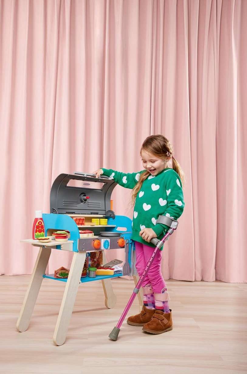 A young girl with a crutch plays with a toy barbecue grill in a room with pink curtains, wearing a green heart-patterned sweater.