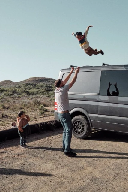 A man throws a child in the air next to a van, with another child watching, on a bright day with sparse desert vegetation in the background.
