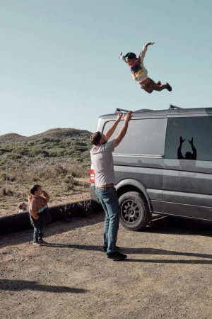 A man throws a child in the air next to a van, with another child watching, on a bright day with sparse desert vegetation in the background.