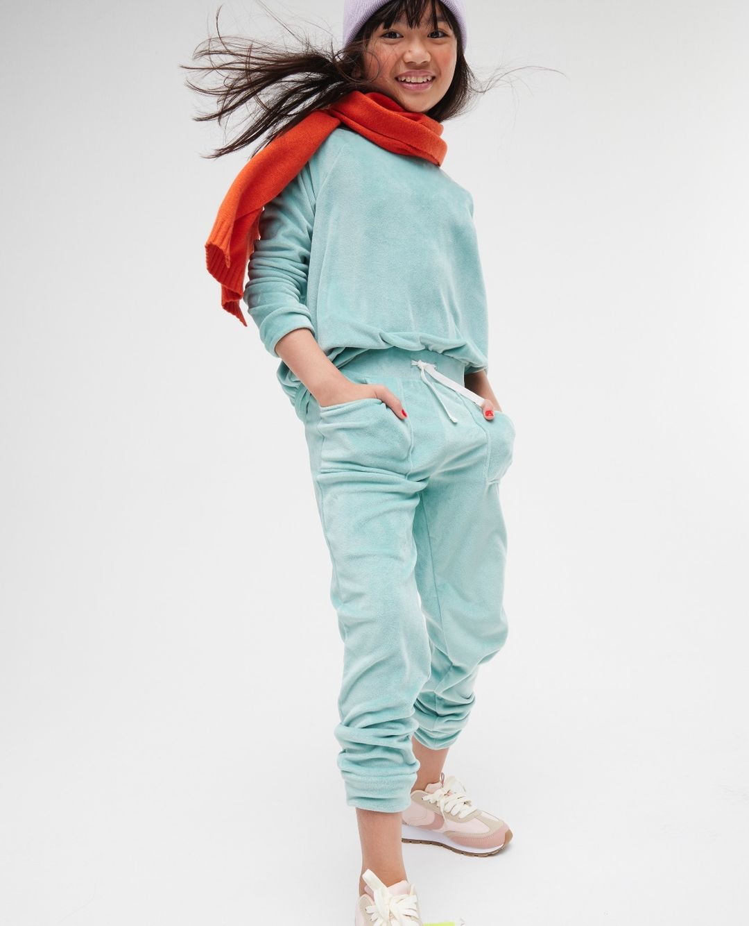 Full shot of a smiling child in a light blue sweat suit, lilac beanie and orange scarf standing against a white backdrop.
