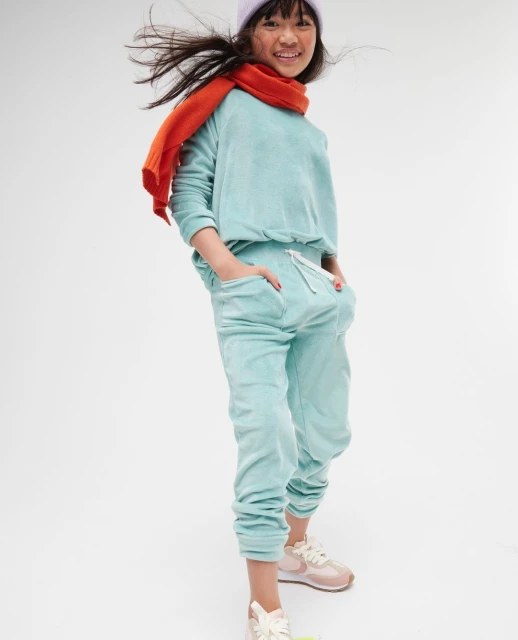 Full shot of a smiling child in a light blue sweat suit, lilac beanie and orange scarf standing against a white backdrop.