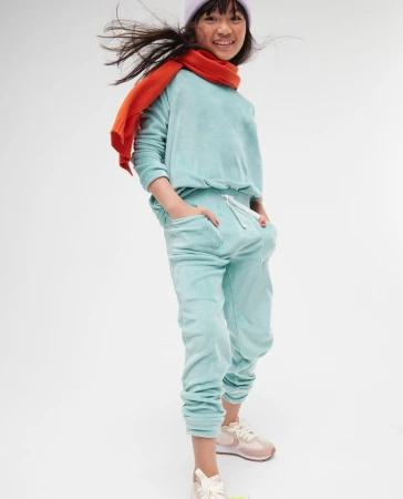 Full shot of a smiling child in a light blue sweat suit, lilac beanie and orange scarf standing against a white backdrop.