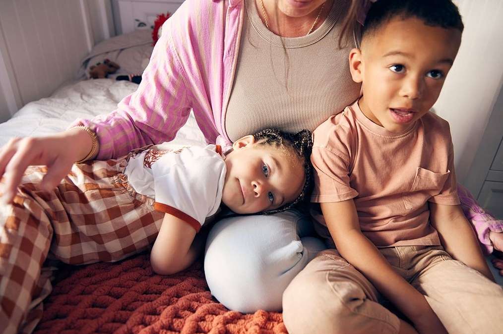 Indoor family scene: a Caucasian woman sits on a bed with two biracial children, one lying on her lap, the other sitting beside her.