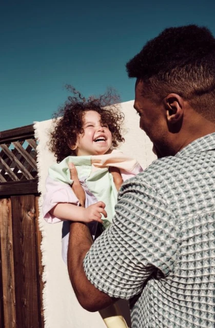 Smiling child with curly hair held up by an adult male in natural light, with a neutral textured background.