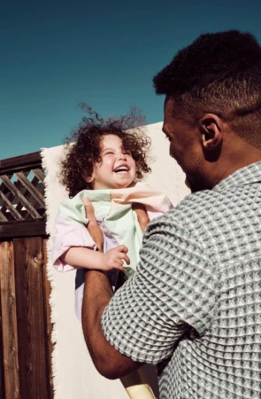 Smiling child with curly hair held up by an adult male in natural light, with a neutral textured background.
