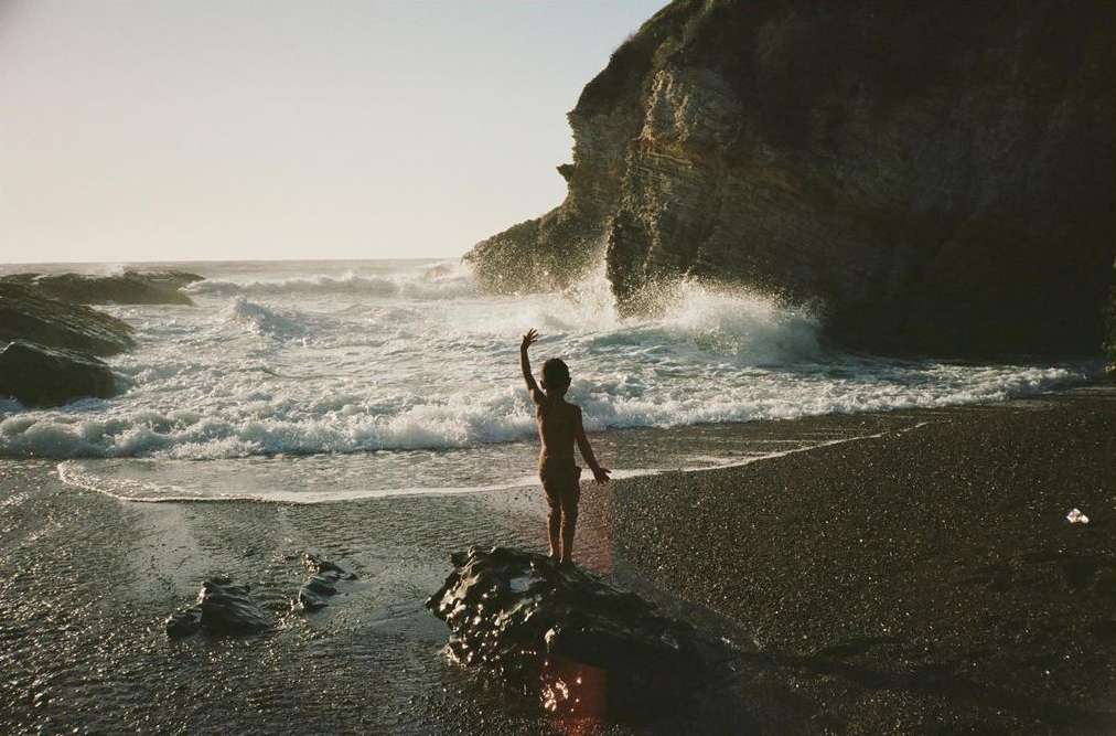 A child stands on a rock at a beach with waves crashing against a rock formation during golden hour for a nature and lifestyle image.