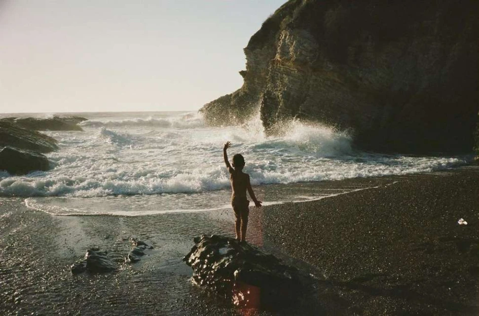 A child stands on a rock at a beach with waves crashing against a rock formation during golden hour for a nature and lifestyle image.