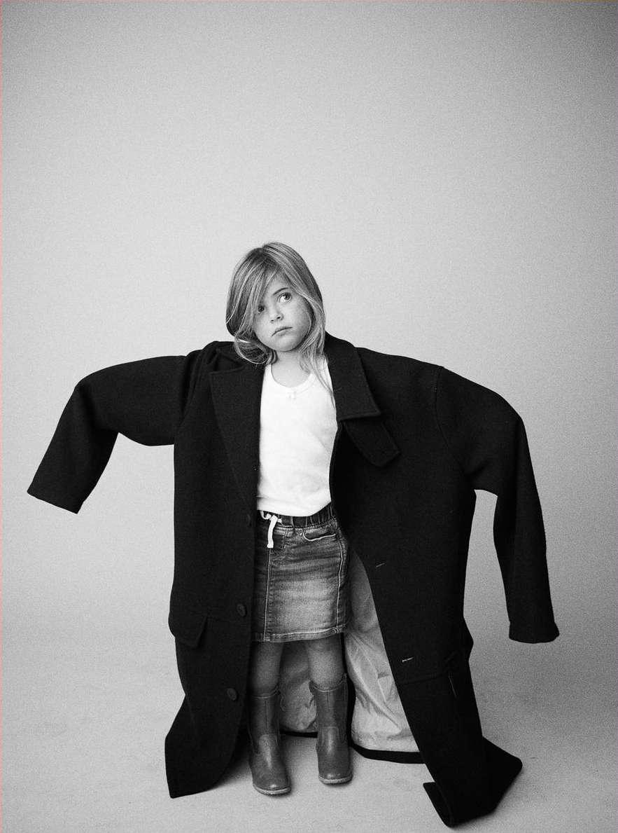 Young girl wearing oversized black coat, posing in photo studio with light gray background. Denim skirt, boots, and white top.