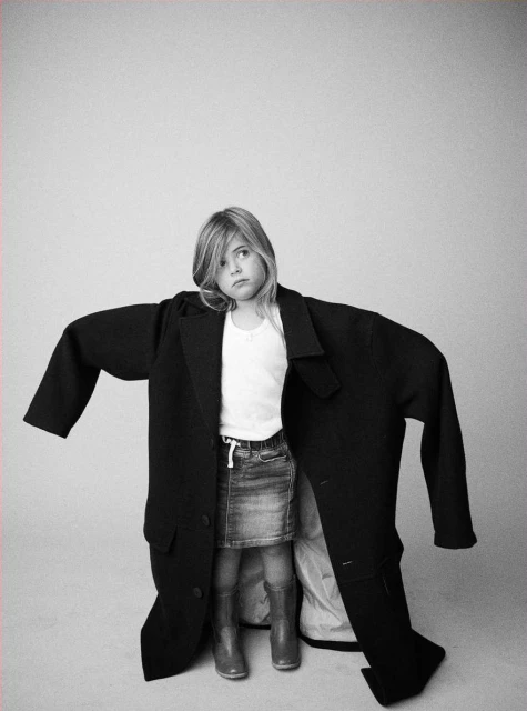 Young girl wearing oversized black coat, posing in photo studio with light gray background. Denim skirt, boots, and white top.
