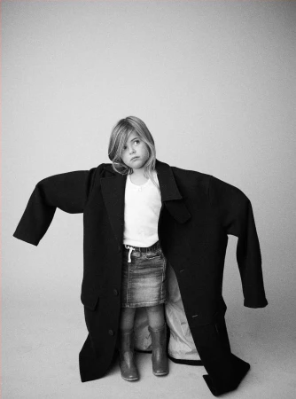 Young girl wearing oversized black coat, posing in photo studio with light gray background. Denim skirt, boots, and white top.