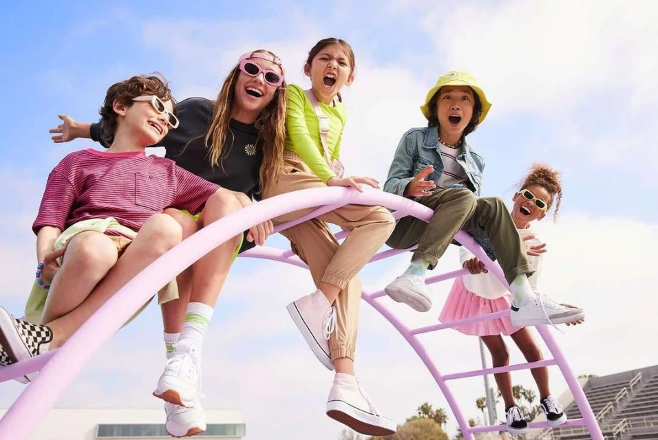 A group of five kids, likely friends, are sitting on a lavender jungle gym with a cloudy blue sky in the background; happy and energetic.