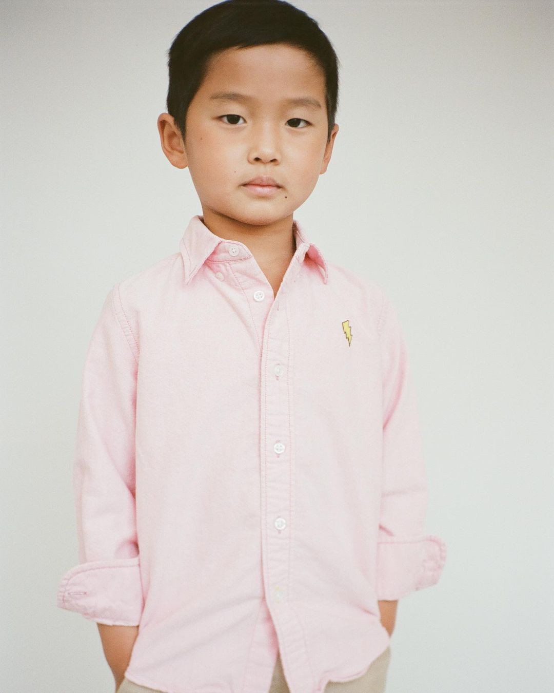 Portrait of an Asian child wearing a light pink button-down shirt with a gold lightning bolt and khaki pants against a white background.