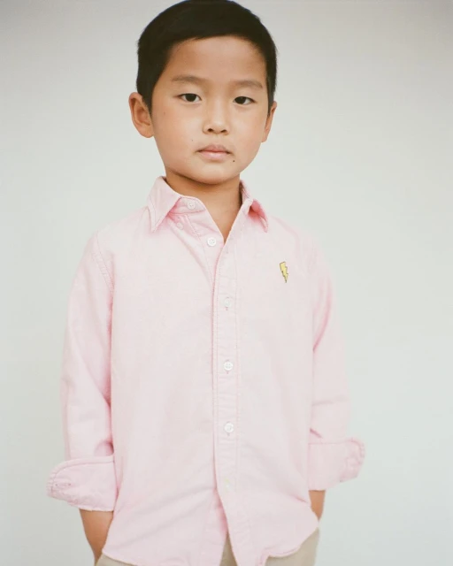 Portrait of an Asian child wearing a light pink button-down shirt with a gold lightning bolt and khaki pants against a white background.