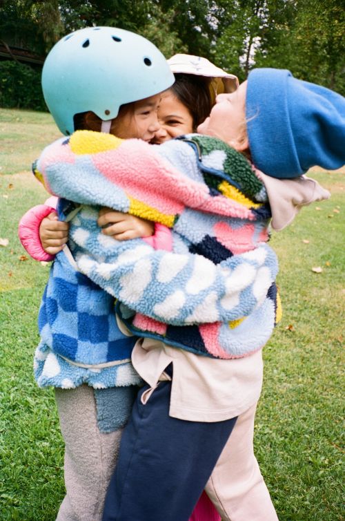 Three children embrace outdoors, wearing colorful jackets and hats, with a light green helmet, in a medium shot with blurred trees.