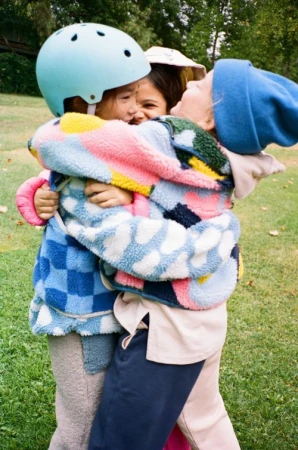 Three children embrace outdoors, wearing colorful jackets and hats, with a light green helmet, in a medium shot with blurred trees.