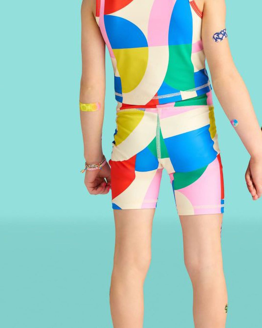 Child in colorful patterned tank top and bike shorts with patches on arms, back view, bright blue background, fashion and kids
