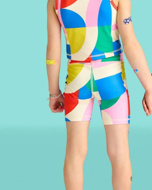 Child in colorful patterned tank top and bike shorts with patches on arms, back view, bright blue background, fashion and kids