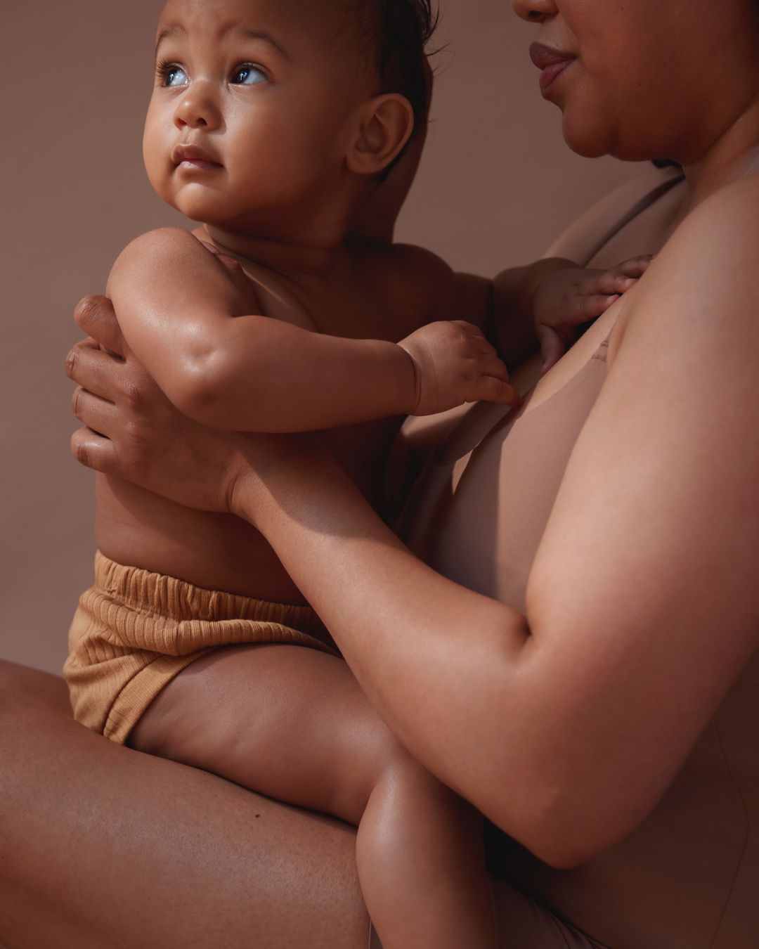 A baby sits in its mother's lap, both wearing tan-colored clothing, close-up, neutral studio lighting, soft focus, and warm skin tones.