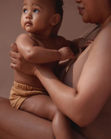 A baby sits in its mother's lap, both wearing tan-colored clothing, close-up, neutral studio lighting, soft focus, and warm skin tones.