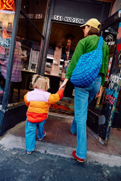 An adult and a child walk hand-in-hand into a building. The adult wears a blue bag, jeans, and a baseball cap. Captured in natural light.