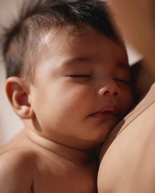 Close-up of a sleeping baby held by an adult with light skin, soft natural light illuminates the serene scene.