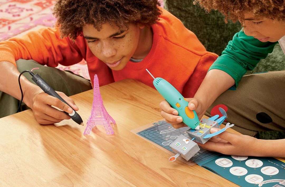 Two children engage in 3D pen art, crafting models on a wooden table with colorful pens. An Eiffel Tower model stands nearby.