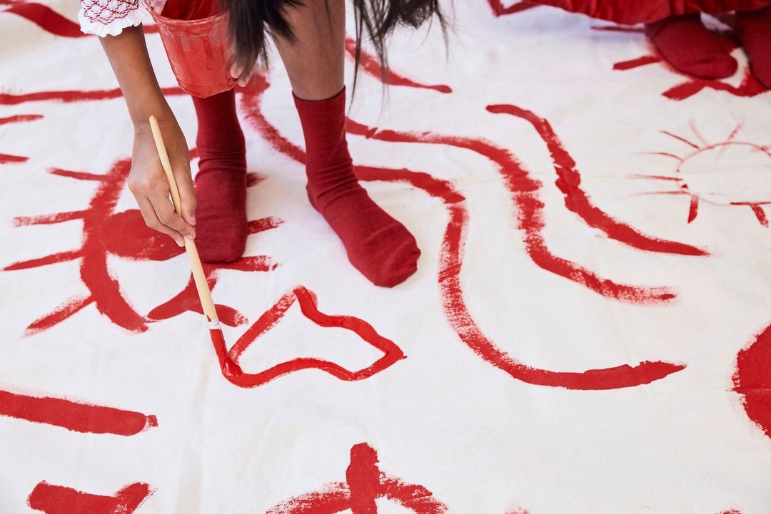 Child creates abstract art with red paint and brush on a white canvas. Visible are the child's arms, legs, hands, and painting tools.