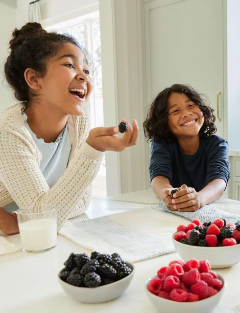 Two smiling children in a brightly lit kitchen at a table with bowls of raspberries and blackberries, plus a glass of milk.