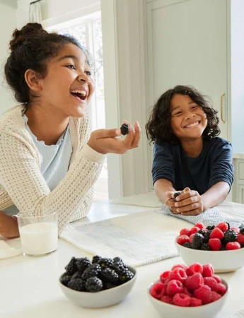Two smiling children in a brightly lit kitchen at a table with bowls of raspberries and blackberries, plus a glass of milk.
