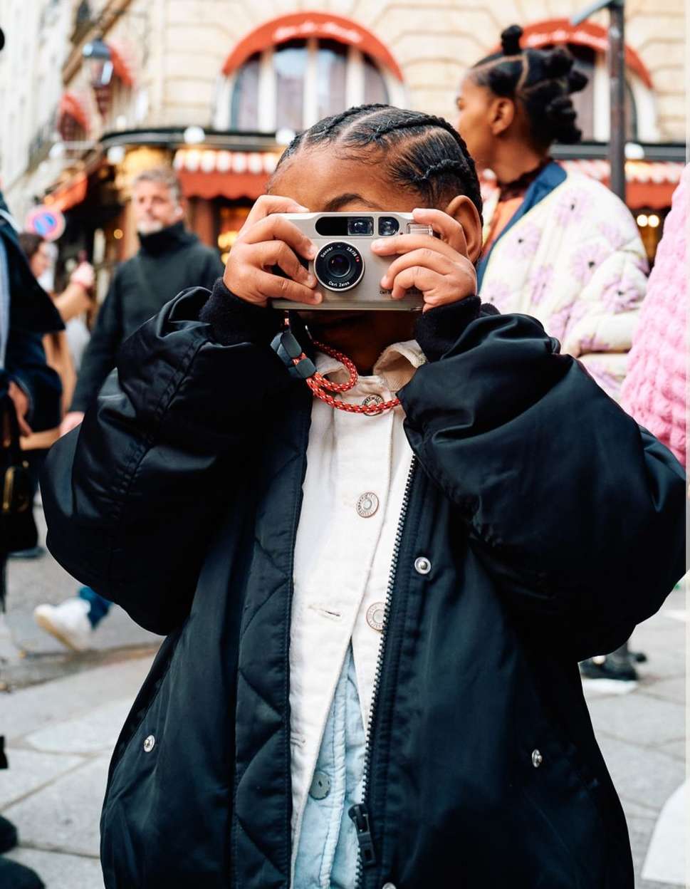 A child holds a camera up to their face in an urban environment with pedestrians in the background, creating a candid street-style photograph.