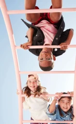 Three children hanging upside down and right side up on a pink ladder against a clear blue sky