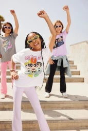 Three young girls posing outdoors on concrete steps in bright daylight wearing graphic t-shirts, sunglasses, and trousers.