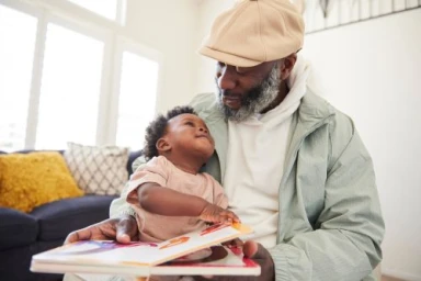 Older Black man wearing a flat cap holding and reading a picture book with a baby sitting on his lap indoors