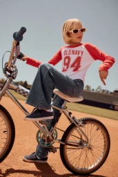 Child with blonde hair wearing sunglasses and a red-and-white raglan t-shirt seated on a vintage bicycle outdoors on a dirt field.