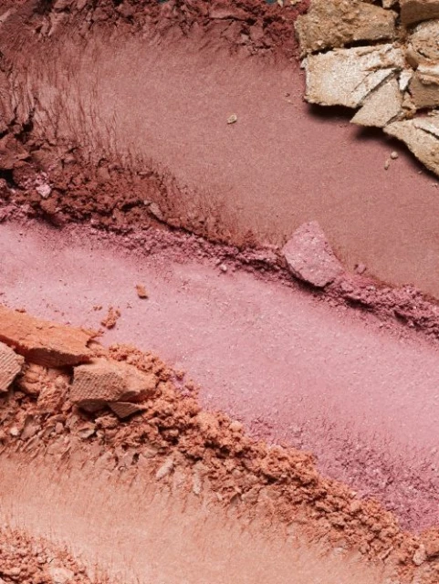 Macro photograph of various shades of powdered makeup including pinks, golds, and browns, swirled together on a neutral surface.