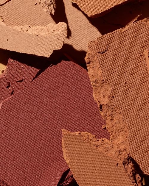 Close-up of shattered makeup powder in warm earth tones including terracotta, umber, sand, and tan creating a textured surface.