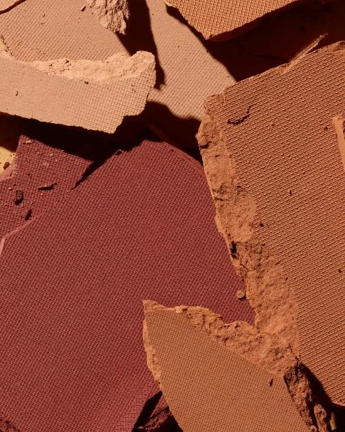 Close-up of shattered makeup powder in warm earth tones including terracotta, umber, sand, and tan creating a textured surface.