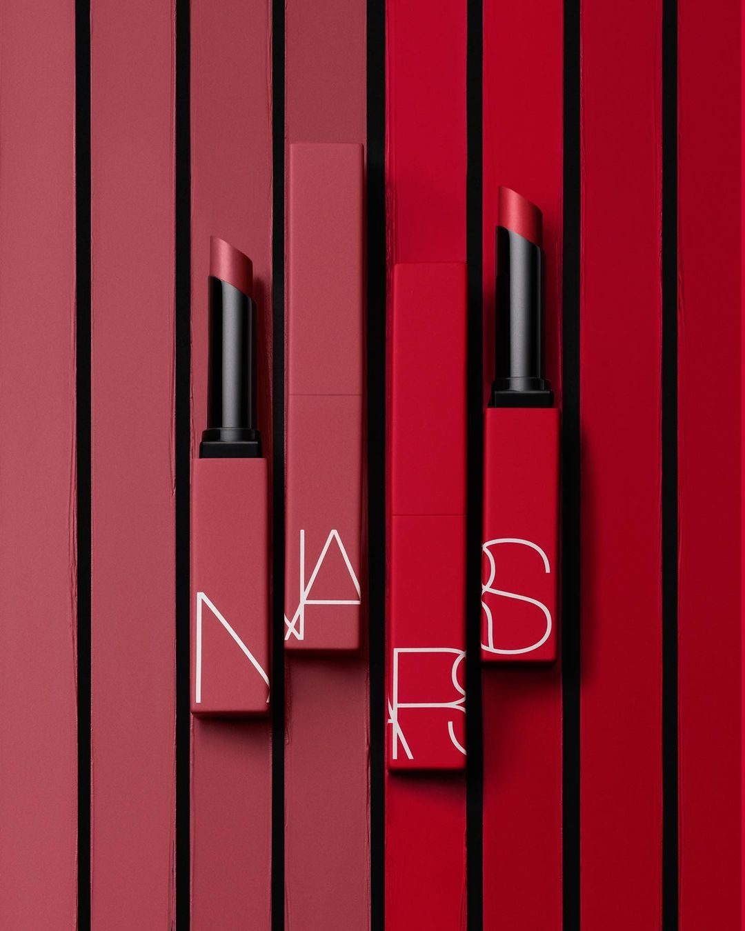NARS lipstick products are arranged against a red and pink striped background in a beauty and cosmetics product still life.