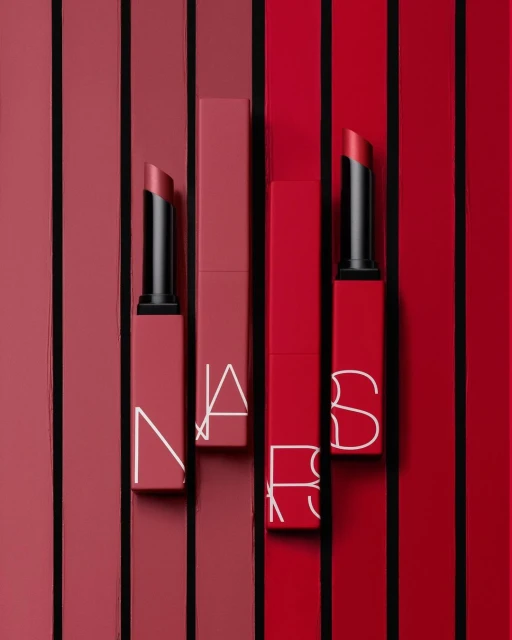 NARS lipstick products are arranged against a red and pink striped background in a beauty and cosmetics product still life.