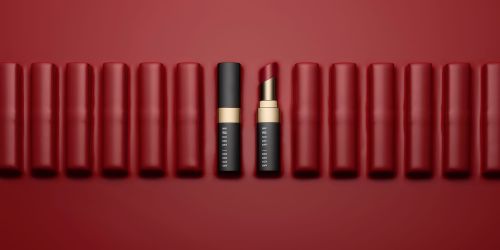 Beauty product shot of arranged red lipstick tubes and one partially open lipstick with gold trim on a red background.
