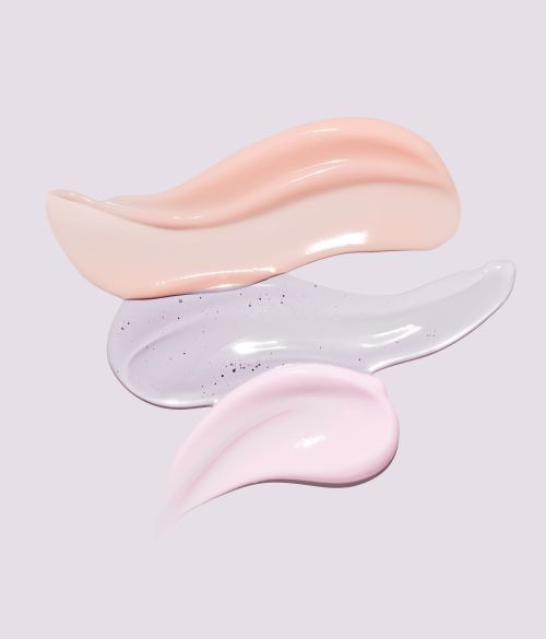 Close-up of three different cosmetic cream smears in peach, light purple with glitter, and light pink against a pale purple background.