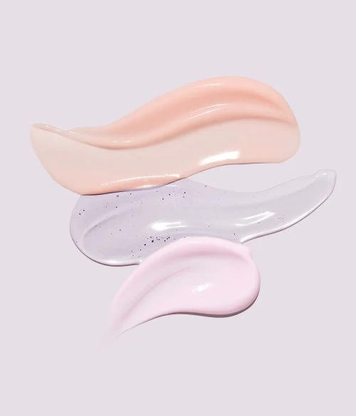 Close-up of three different cosmetic cream smears in peach, light purple with glitter, and light pink against a pale purple background.