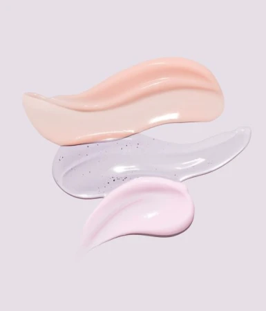 Close-up of three different cosmetic cream smears in peach, light purple with glitter, and light pink against a pale purple background.