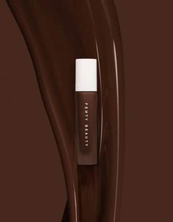 Fenty Beauty liquid cosmetic product bottle positioned vertically against a flowing rich brown liquid backdrop